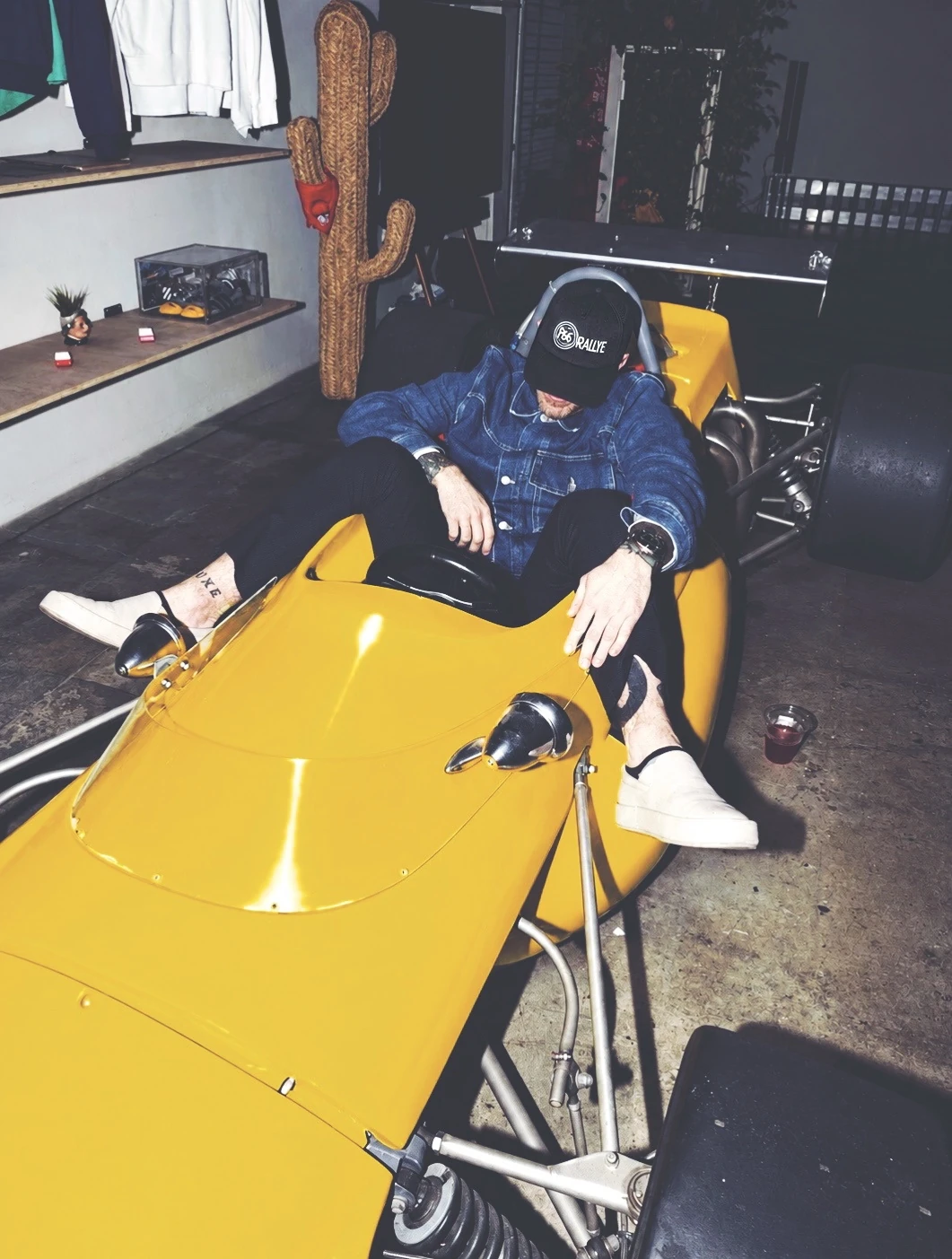 A man in a denim jacket and a P66 Rallye cap lying back across the nose of a yellow single-seater in a shop at night